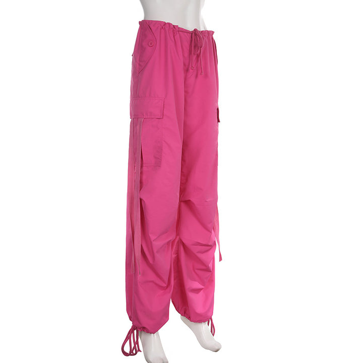 Un mannequin porte un pantalon rose baggy taille basse pour femme, avec une coupe large façon parachute et des lanières pendantes. Confortable et extensible, ce pantalon cargo est disponible en tailles S à L.