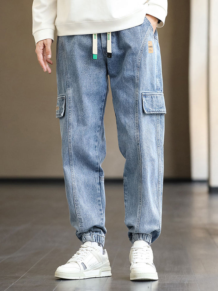 Un homme portant un jean cargo jogger bleu avec élastique à la taille et cordon bicolore blanc et turquoise. Coupe légèrement large, patchs marron et coutures structurantes ajoutant une touche tendance. Disponible du M au 8XL.