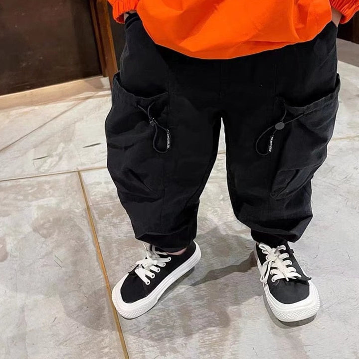 Un enfant pose dans une pièce avec mur marron et carrelage gris au sol. Il porte un pull sweat orange, des baskets noires et blanches ainsi qu'un pantalon cargo noir avec de grandes poches latérales.