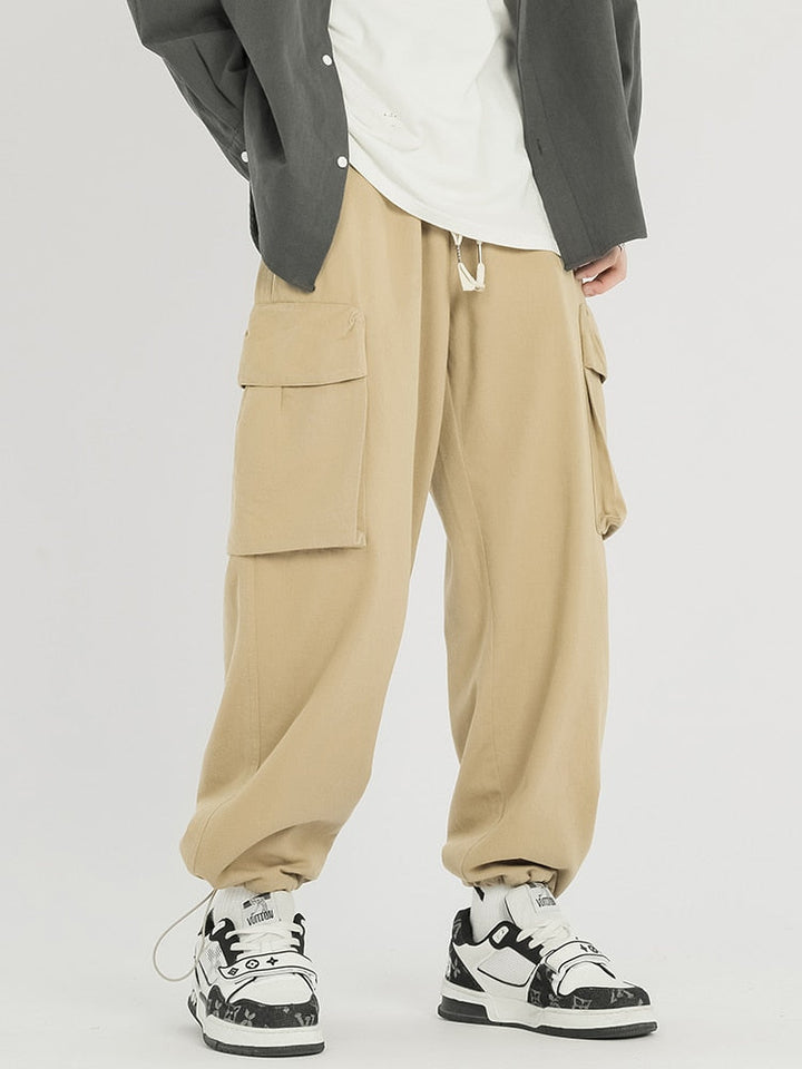 Un homme pose en pantalon cargo baggy en coton beige. Il porte également un tee shirt blanc, une chemise ouverte grise et des baskets noires et blanches.