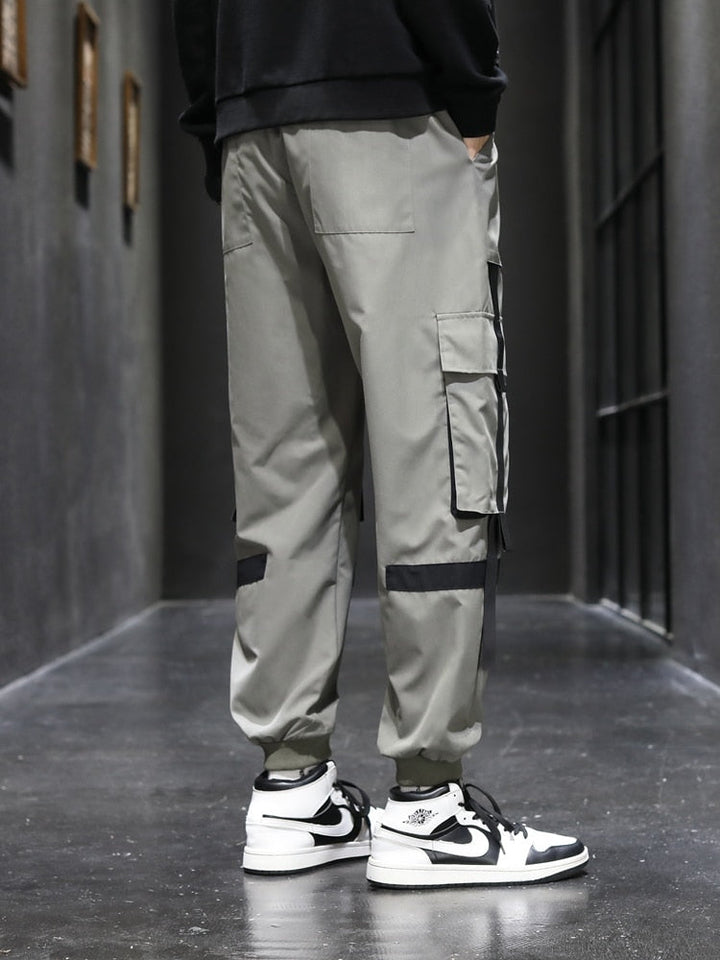 Un homme porte un pantalon cargo kaki avec des lanières resserrées aux chevilles. Le style street masculin est accentué par des lanières noires sur les poches et les jambes. Disponible du M au 5XL.