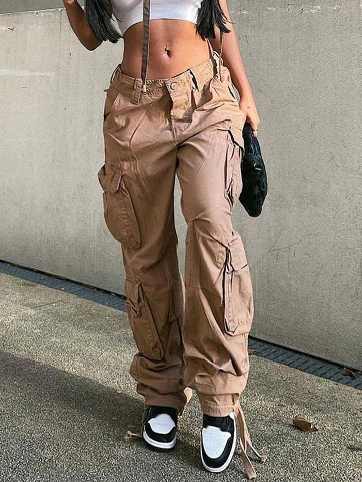 Une femme pose dans la rue avec un crop top blanc, des baskets noires et blanches ainsi qu'un pantalon cargo coupe droite avec de nombreuses poches camel. Elle porte un sac noir et a un piercing au nombril et des cheveux longs bruns.