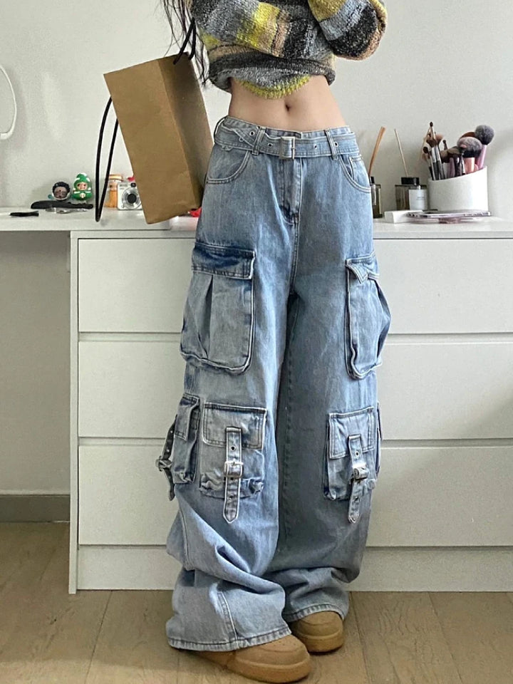 Une femme pose dans sa chambre en jean baggy cargo. Le jean a de nombreuses poches et il est taille basse avec une ceinture. 