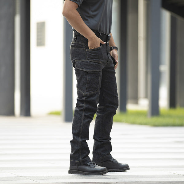 Un homme pose dans la rue avec un jean cargo ajusté coupe droite noir, un tee-shirt et des boots noirs. Il a une ceinture et une montre noires et porte son téléphone à la main.