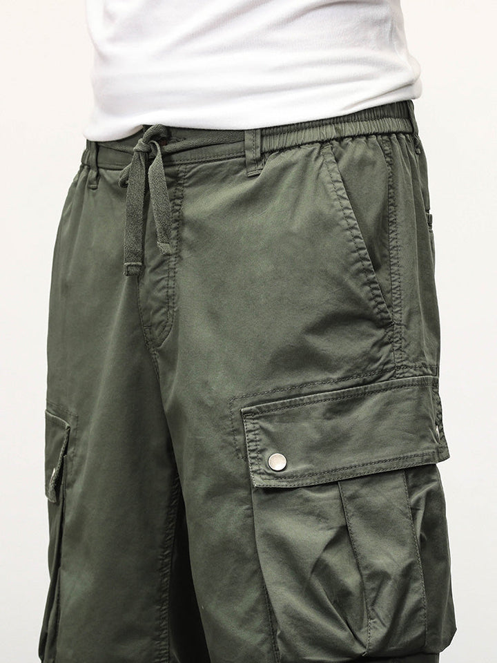 Un homme portant un short cargo kaki coupe droite avec des poches latérales à boutons-pression. Fabriqué à partir de 95% coton et 5% spandex, ce short offre confort et élasticité. Tailles disponibles du 28 au 40.