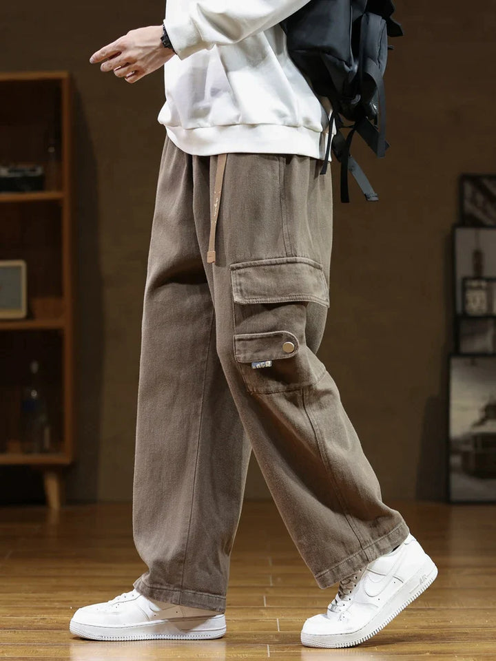 Un homme porte un pantalon cargo ample en coton marron de notre boutique Cargo District. Ce pantalon résistant offre une grande liberté de mouvement grâce à sa coupe ample. La ceinture intégrée ajoute une touche utilitaire chic. Disponible en différentes tailles.