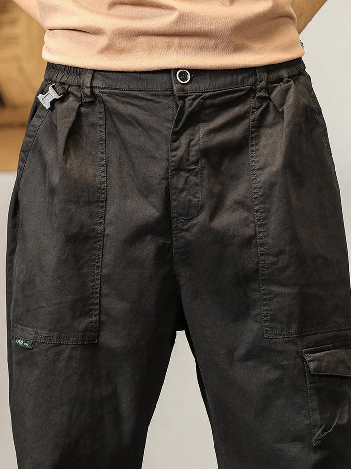 Un homme porte un pantalon cargo noir ample coupe droite avec des poches fonctionnelles. Matière légère et confortable. Tailles disponibles du M au 3XL. (Cargo District)