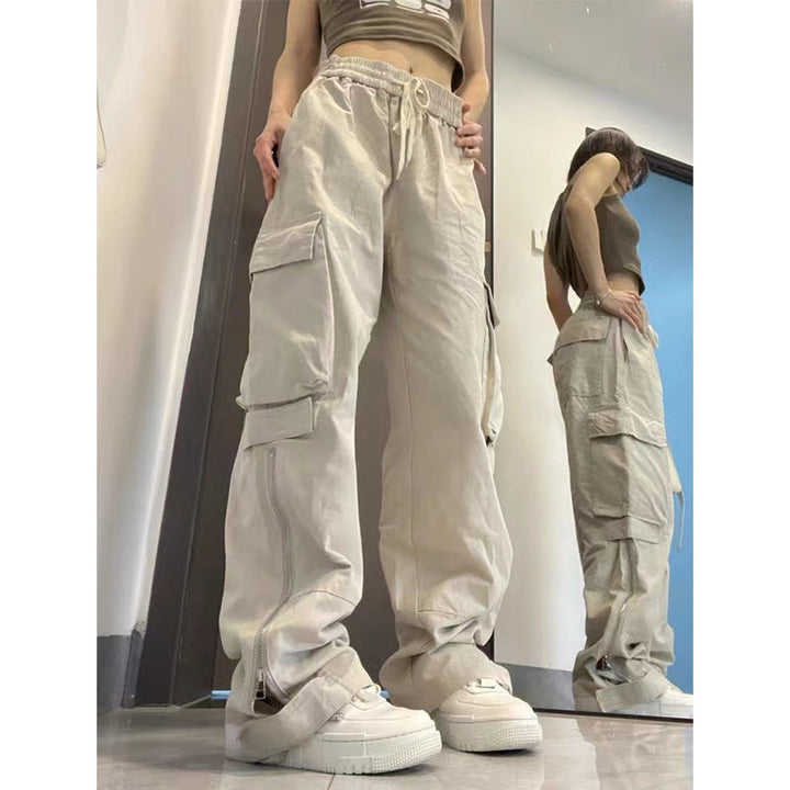 Une femme pose en pantalon baggy avec fermeture éclair sur le bas de la jambe. Le baggy est beige. Il porte des baskets blanches, un crop top taupe. 