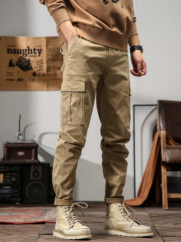 Un homme pose dans un décor industriel avec un sweat camel, une montre et des boots marrons. Il porte également un pantalon cargo coupe droite beige.