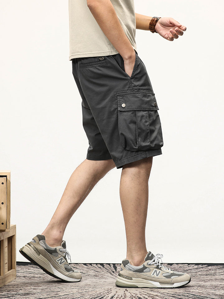 Un homme portant un short cargo gris coupe droite et des baskets. Le short cargo offre une coupe ample et des poches latérales à boutons-pression pour une touche pratique. Tailles disponibles du 28 au 40. Confortable et élastique grâce à sa composition en 95% coton et 5% spandex. Parfait pour adopter un style cargo intemporel.