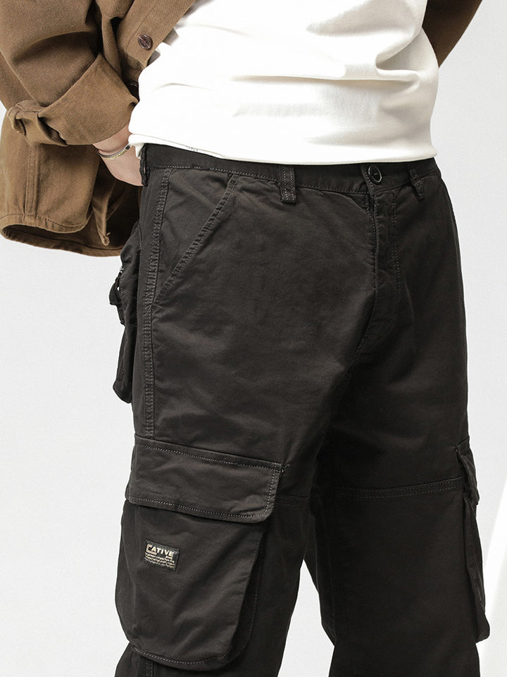 Un homme porte un pantalon cargo noir ample coupe droite avec poches latérales. Fermeture par bouton et fermeture éclair. Allie fonctionnalité et style. Jambe large resserrable aux chevilles. Disponible en tailles 28 à 38 (US). Matières : 97% coton, 3% Spandex pour confort et flexibilité.