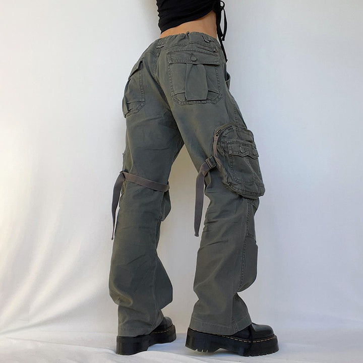 Une personne portant un pantalon cargo large avec des lanières autour des cuisses, couleur kaki, pour femme. Les lanières cousues aux poches latérales ajoutent une touche militaire. Disponible du S au L.