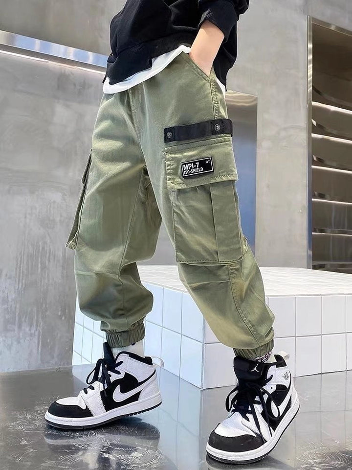 Un enfant porte un pantalon cargo jogger large kaki avec des baskets noires. Le pantalon a des poches latérales pratiques et une taille élastique pour un ajustement parfait. Disponible en tailles de 110 à 160 cm. Style urbain et confortable.