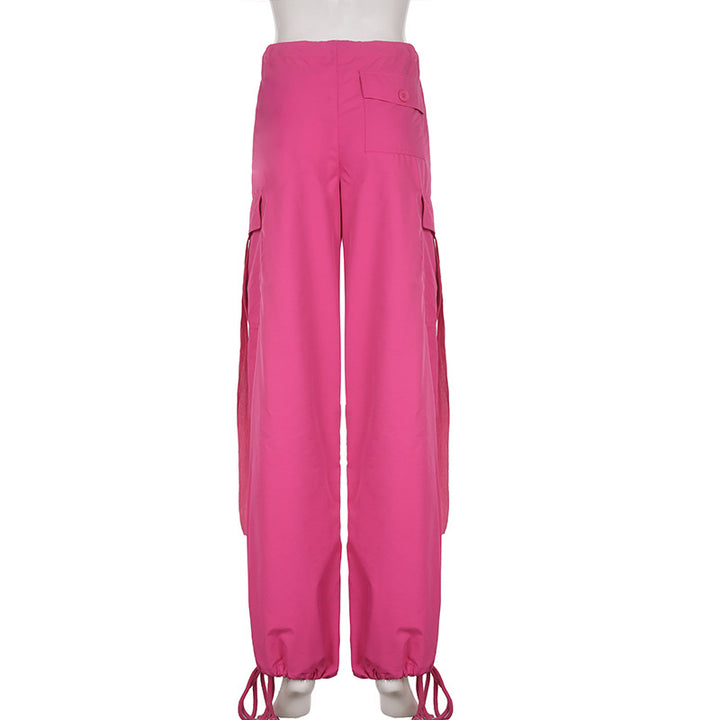 Un mannequin porte un pantalon cargo baggy taille basse rose pour femme avec une coupe large façon parachute et des lanières pendantes. Confortable et extensible, ce pantalon est disponible en tailles S à L. Parfait pour un style street féminin audacieux.