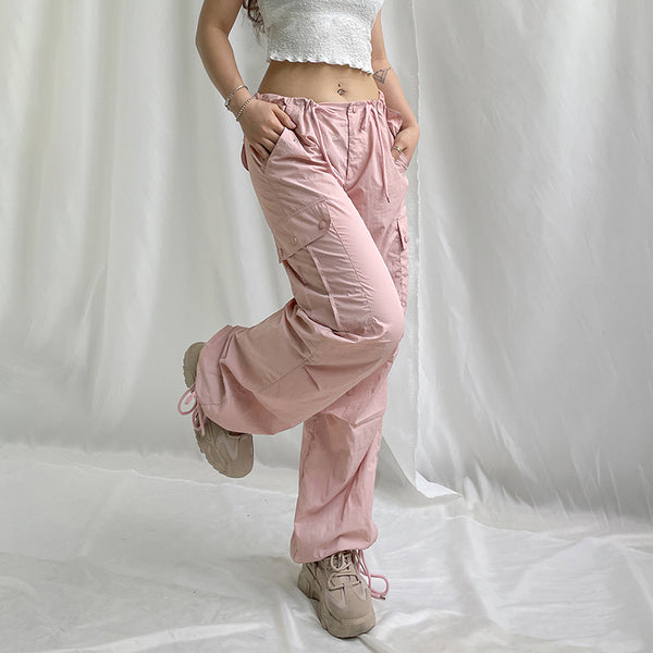 Une femme pose avec devant un fond blanc un tissu. Elle pose avec tee-shirt blanc court, un pantalon cargo taille basse large rose avec élastique aux chevilles. Elle porte aussi des baskets marron clair. 