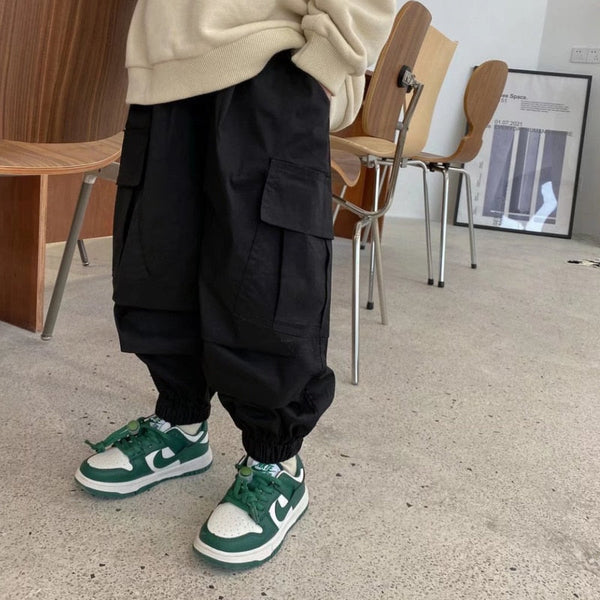 Un enfant pose dans un salon aux murs blancs sol beige avec des chaises en bois. L'enfant porte des baskets blanches et vertes, un pantalon cargo noir baggy, un sweat beige. 