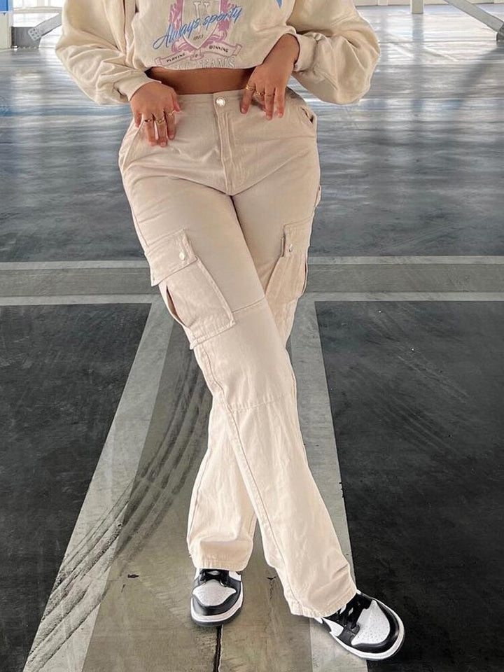 Une femme porte un pantalon cargo ajusté taille haute beige. Ce pantalon combine style et fonctionnalité avec sa coupe ajustée qui s'évase légèrement sur le bas. Confectionné en coton, il offre un confort ultime et une durabilité exceptionnelle. Disponible du S au XL.