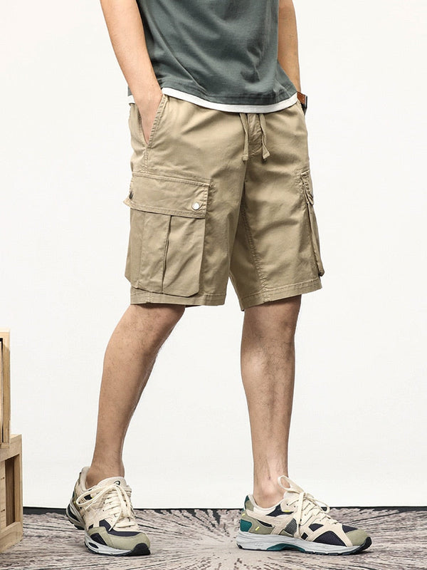 Un homme pose dans une pièces avec tapis gris au sol, mur blanc et des caisses en bois sont sur le côté de la photo. Il porte des baskets, un tee-shirt kaki et un short cargo coupe droite beige.