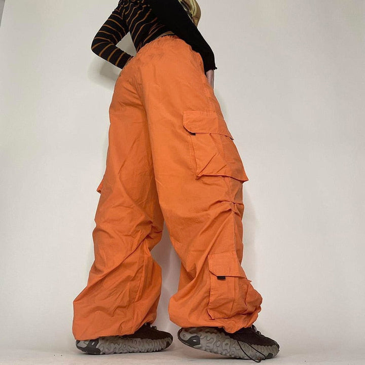 Une femme portant un pantalon orange baggy taille basse avec des détails de lanières pendantes, créant une silhouette unique et audacieuse. Le pantalon cargo baggy taille basse est disponible du S au L et offre un confort sans égal grâce à sa coupe large façon parachute et sa texture douce et extensible en polyester, jersey et spandex. Les chevilles élastiques ajoutent à l'effet baggy. Un choix parfait pour le style street féminin.