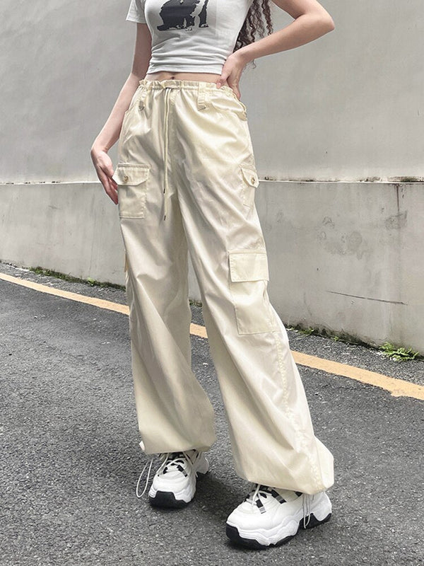 Une femme pose dans la rue avec un pantalon baggy taille basse blanc cassé, avec taille élastique. Elle porte aussi un tee-shirt beige et des baskets blanches. 