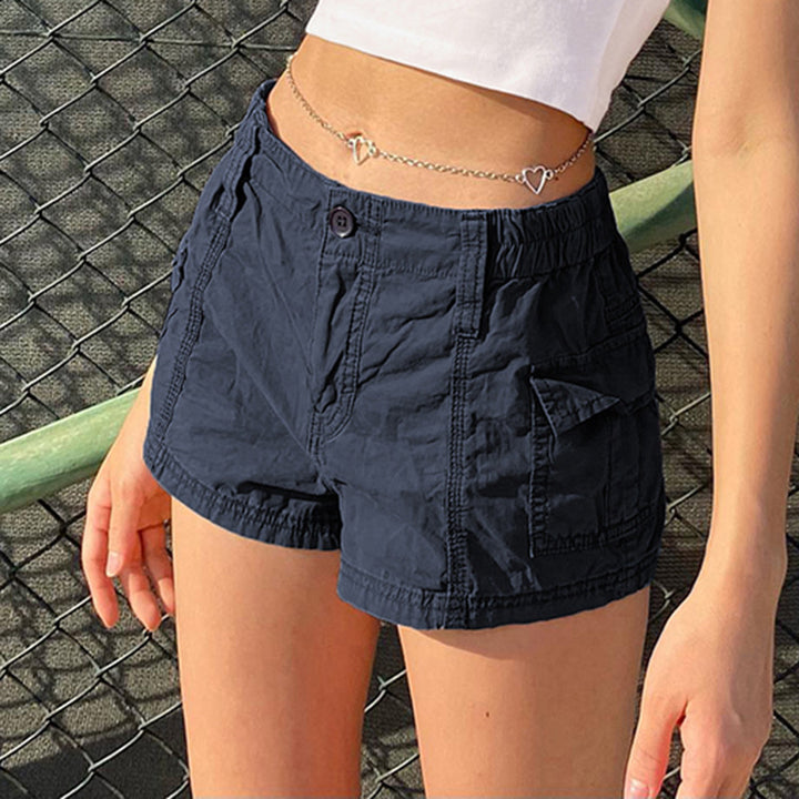 Une femme pose en short cargo bleu, avec un crop top blanc et derrière un grillage. Elle porte une chaîne de ventre argenté avec coeurs.