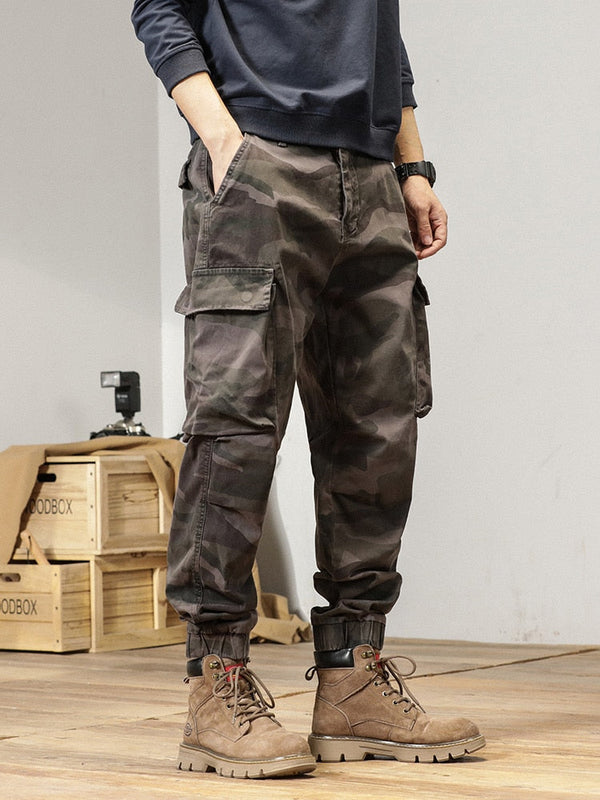 Un homme pose dans une pièce avec parquet au sol et murs blancs. Il y a des boîtes en bois et un appareil photo derrière lui. Il porte un sweat gris, des boots marrons et un pantalon cargo ample resserré aux cheviles gris camo.