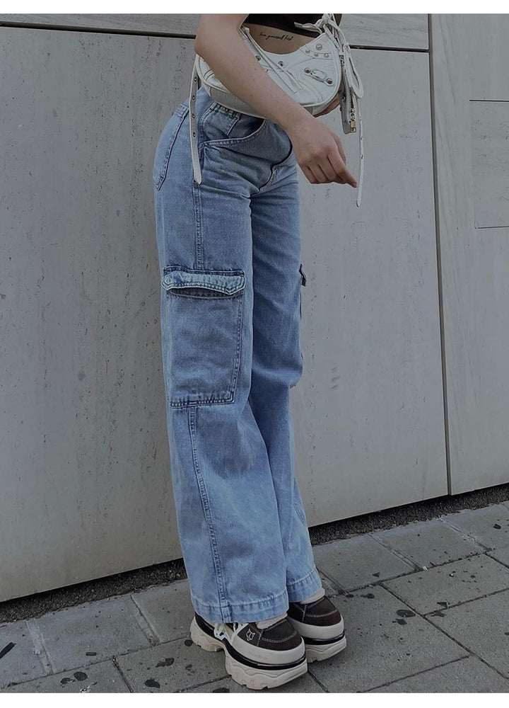 Une personne debout à côté d'un mur, portant un jean cargo coupe droite taille haute bleu pour femme. Les poches latérales cargo ajoutent une touche distinctive et urbaine à ce pantalon. Disponible du S au XL.