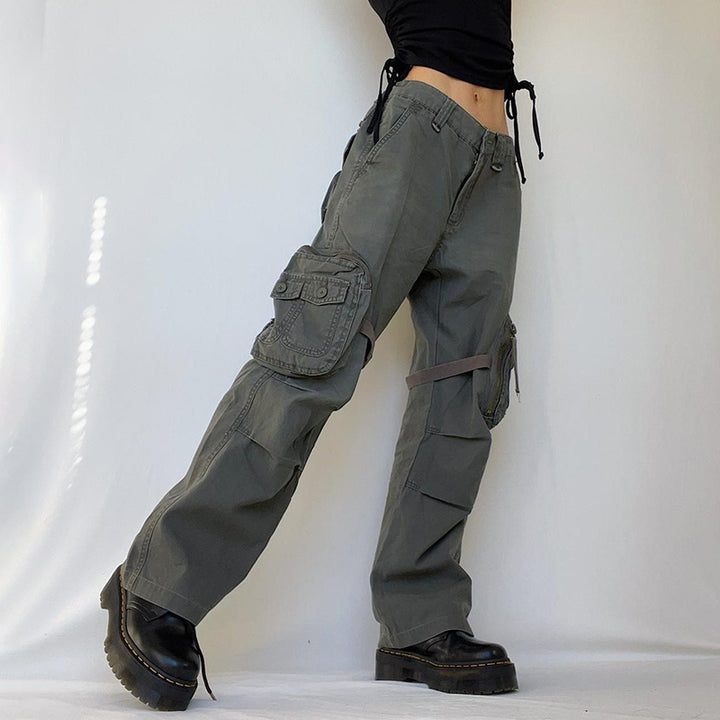 Une personne porte un pantalon cargo large avec des lanières autour des cuisses, de couleur kaki. Les poches latérales sont ornées de lanières qui s'enroulent autour de la jambe, ajoutant une touche militaire. Ce pantalon cargo est idéal pour un look urbain audacieux.