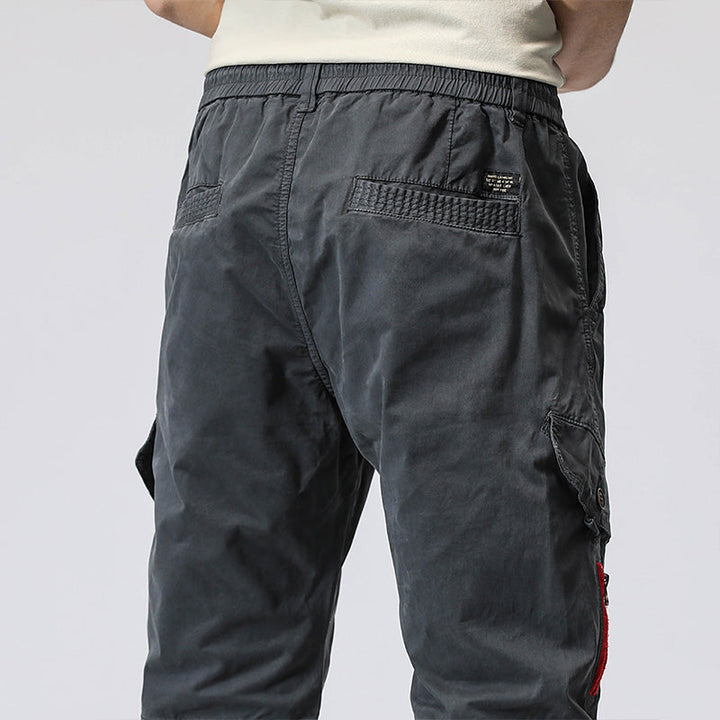 Un homme porte un pantalon cargo gris coupe droite avec fermeture rouge. Sa coupe resserrée à la cheville structure la silhouette. Disponible du 28 au 38 (tailles US).