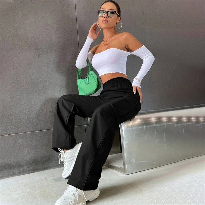 Une femme assise sur un banc, portant le Pantalon Cargo Baggy noir pour femme de Cargo District. Le pantalon taille haute en polyester léger et durable met en valeur la taille et les courbes féminines. Les jambes larges et les multiples poches ajoutent une touche streetwear et fonctionnelle. Disponible du S au XL.