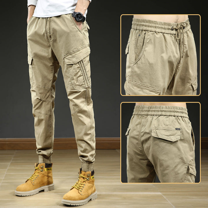 Un homme porte un pantalon cargo slim resserré aux chevilles de couleur beige. Le pantalon est confortable et élégant, avec une coupe slim et des coutures aux genoux. Disponible en tailles US 28 à 38.