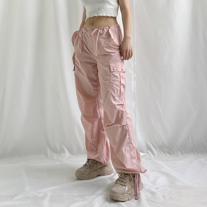 Une femme portant un pantalon cargo rose avec une taille basse et une jambe large. Le pantalon est fini par un élastique à la cheville pour un style street. Disponible du S au L. Matières : Polyester, Spandex. Confortable et urbain.