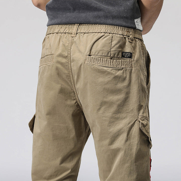 Un homme porte un pantalon cargo coupe droite avec fermeture rouge, de couleur vert olive. La coupe droite et resserrée à la cheville structure sa silhouette. Le pantalon se distingue par une fermeture éclair rouge sur l'une des poches, ajoutant une touche de couleur audacieuse. Disponible dans les tailles 28 à 38 (tailles US), ce pantalon offre confort et style décontracté.