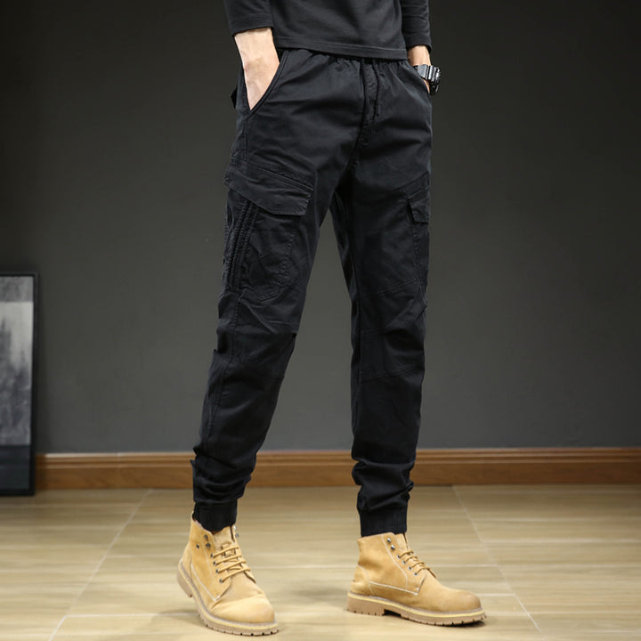 Un homme porte un pantalon cargo slim resserré aux chevilles de couleur noire avec des bottes marron. Le pantalon est confortable et élégant, avec une coupe moderne et des coutures aux genoux. Disponible en tailles US 28 à 38.