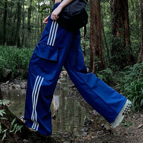 Une femme pose en forêt avec un sac à dos, des baskets blanches et un pantalon baggy bleu à bandes latérales blanches. Le pantalon est taille haute et large.