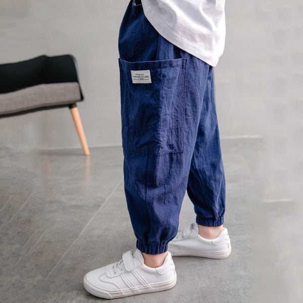 Un enfant pose dans une pièce avec murs et sol gris et un fauteuil noir dans le fond.  Il porte un tee-shirt blanc, des baskets blanches ainsi qu'un pantalon cargo jogger en coton bleu.