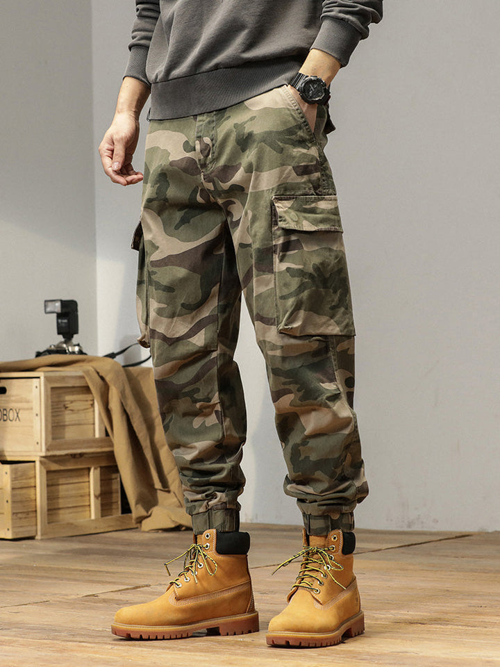 Un homme portant un pantalon cargo kaki, coupe large resserré aux chevilles. Style militaire masculin, confortable et durable en coton 100%. Tailles US 28 à 38.