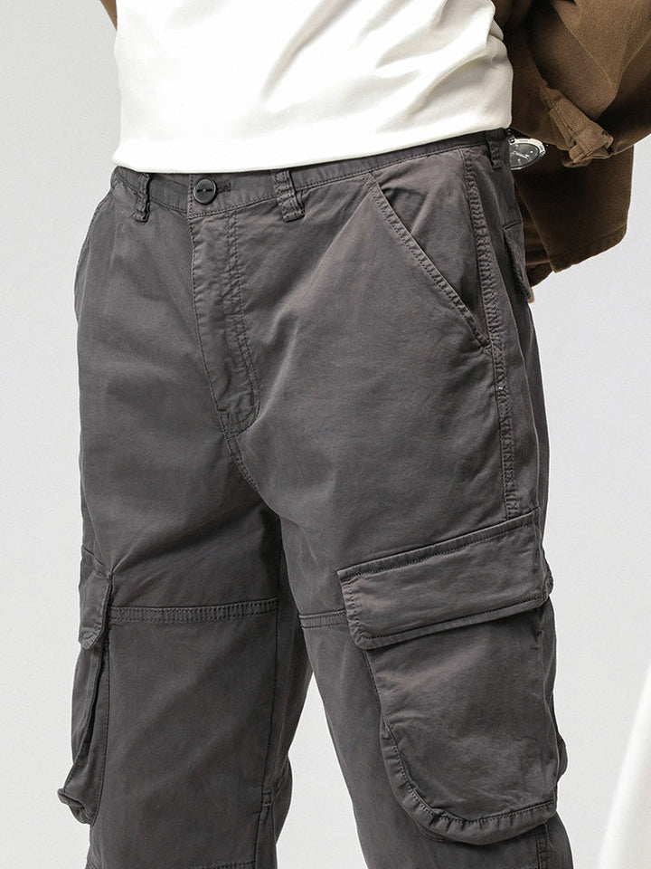 Un homme porte un pantalon cargo ample coupe droite de couleur grise. Le pantalon est en coton et spandex, offrant confort et flexibilité. Il comporte des poches sur les côtés et se ferme par bouton et fermeture éclair. La jambe large peut être resserrée aux chevilles avec un lien. Disponible en tailles du 28 au 38 (US).