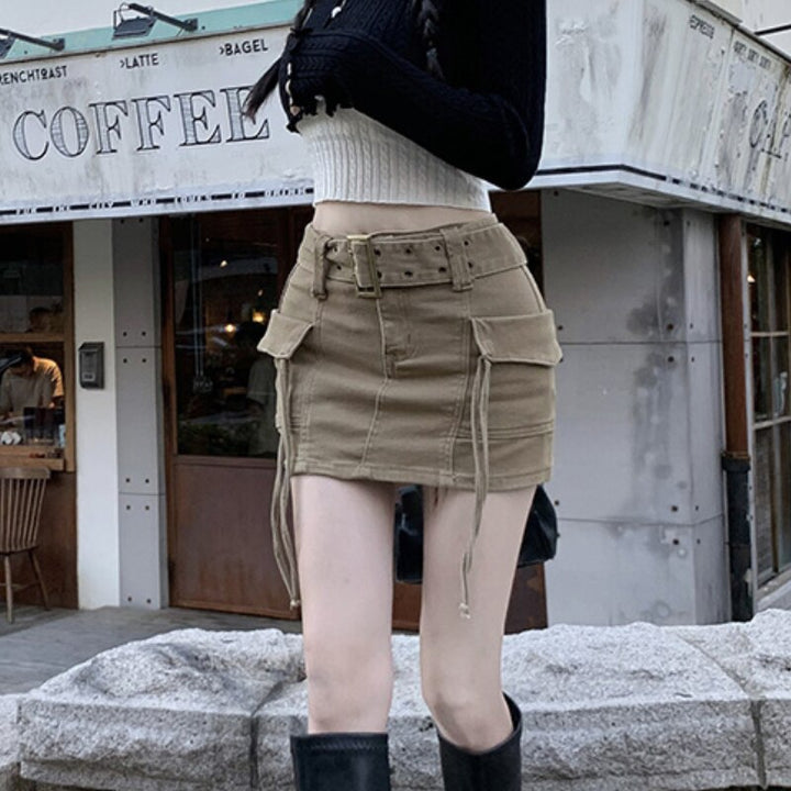 Une femme debout à l'extérieur d'un bâtiment, portant une jupe cargo beige avec ceinture et lanières. Silhouette cintrée et droite, ajoutant une touche féminine à ce style typiquement cargo. Disponible du S au 2XL.