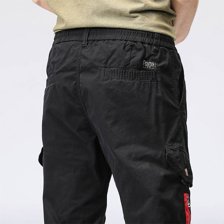 Un homme porte un pantalon cargo noir avec une fermeture éclair rouge sur une poche. Coupe droite et resserrée à la cheville. Disponible en tailles 28-38. Matières : 95% coton, 5% Spandex.