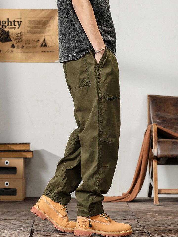 Un homme debout dans une pièce portant un pantalon cargo ample coupe droite kaki. Les poches sur les côtés ajoutent une touche fonctionnelle. Disponible du M au 3XL.