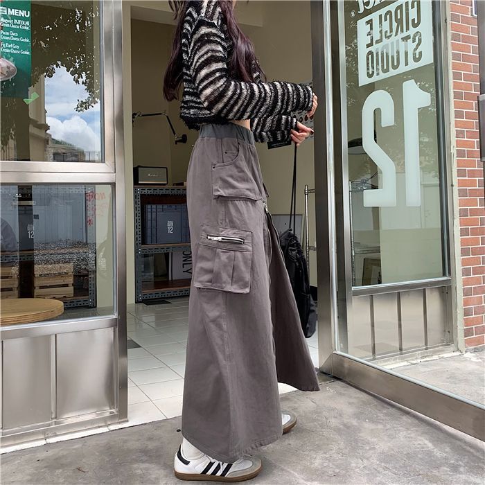 Une femme debout devant un magasin, portant une Jupe cargo longue zippée grise. La jupe a une coupe évasée et une taille haute avec élastique pour plus de confort et de maintien. Une fermeture éclair verticale sur le devant ajoute une touche d'originalité. Disponible en tailles S à XL.