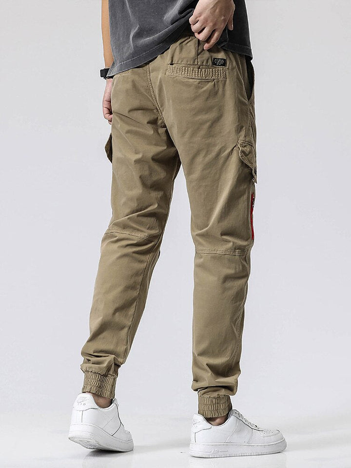 Un homme porte un pantalon cargo coupe droite avec fermeture rouge, de couleur vert olive. Le pantalon est resserré à la cheville et possède une fermeture éclair rouge sur l'une des poches. Disponible dans les tailles 28 à 38 (US). Matières : 95% coton et 5% Spandex. Idéal pour un look élégant et décontracté.