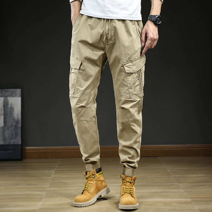 Un homme portant un pantalon cargo slim resserré aux chevilles, couleur beige, avec des bottes jaunes. Le pantalon est confortable et élégant, avec une coupe moderne et des coutures aux genoux. Disponible en tailles US 28 à 38.