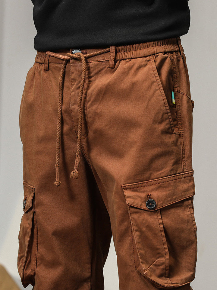 Un homme porte un pantalon cargo coupe droite avec des poches à bouton de couleur camel. Les poches à rabats sont équipées d'un bouton apparent pour une fermeture sécurisée. Le pantalon offre un look masculin avec sa coupe droite et son bas resserré aux chevilles. Disponible en tailles US 28 à 38.