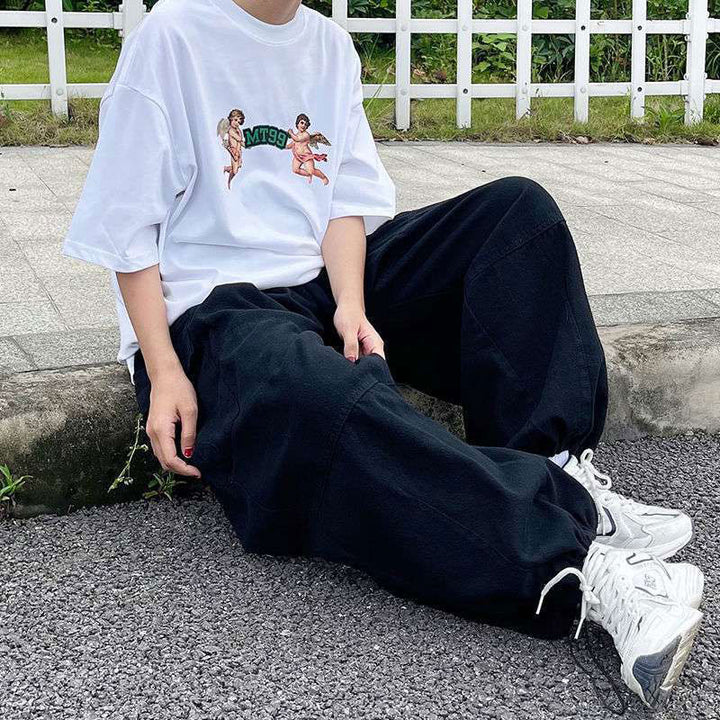 Une femme pose assise sur le trottoir avec un tee-shirt blanc avec des anges, des baskets blanches ainsi qu'un pantalon cargo baggy extra- large noir. 