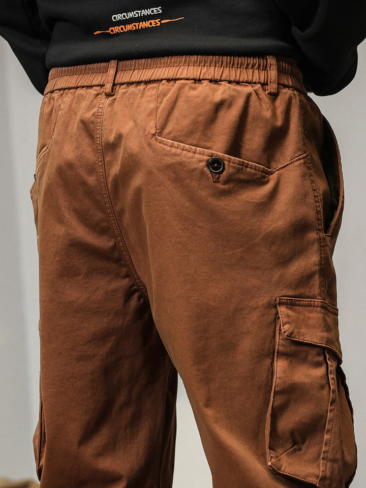 Un homme portant un pantalon cargo coupe droite avec poches à bouton de couleur camel. Le pantalon a des poches à rabats avec des boutons apparents pour une fermeture sécurisée. Il a une coupe droite avec un bas resserré aux chevilles, offrant un look masculin. Tailles disponibles: US 28 à 38.