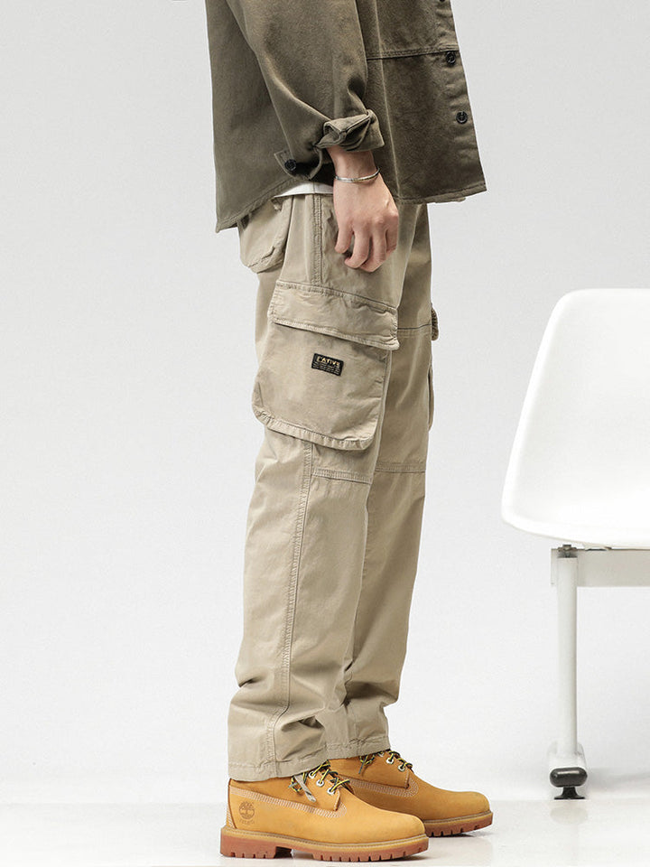 Un homme porte un pantalon cargo ample coupe droite de couleur beige. Le pantalon est pratique avec ses poches sur les côtés et sa fermeture par bouton et fermeture éclair. Il peut être resserré aux chevilles pour un look sportif ou porté droit pour un look classique. Disponible en différentes tailles.