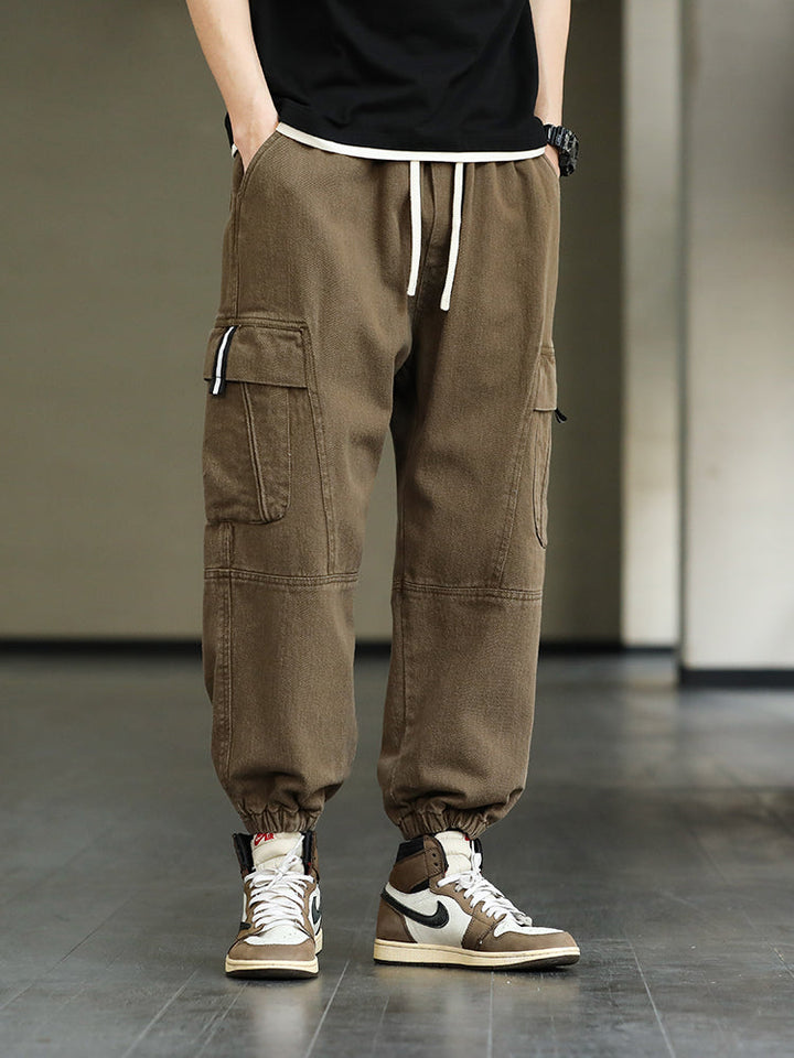 Un homme porte un pantalon cargo ample resserré aux chevilles de couleur marron clair. Les poches cargo latérales ajoutent une touche fonctionnelle. Ce pantalon est disponible en différentes tailles.