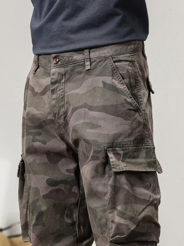 Un homme porte un pantalon cargo coupe large resserré aux chevilles de couleur gris camo, style militaire. Conçu en 100% coton pour un confort optimal et une durabilité à toute épreuve. Disponible en tailles US 28 à 38.